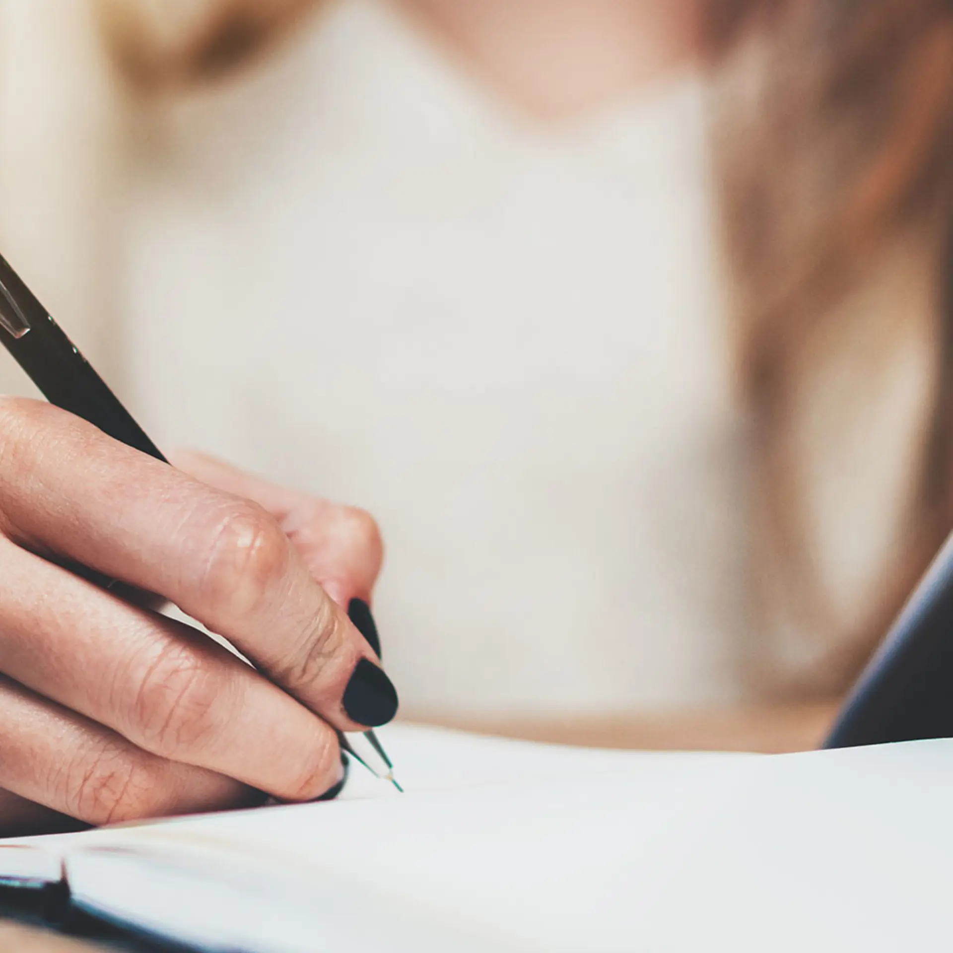 Kvinde med løst hår og sort neglelak skriver i sin notesbog. Med sin venstre hånd holder hun på et dokument. 