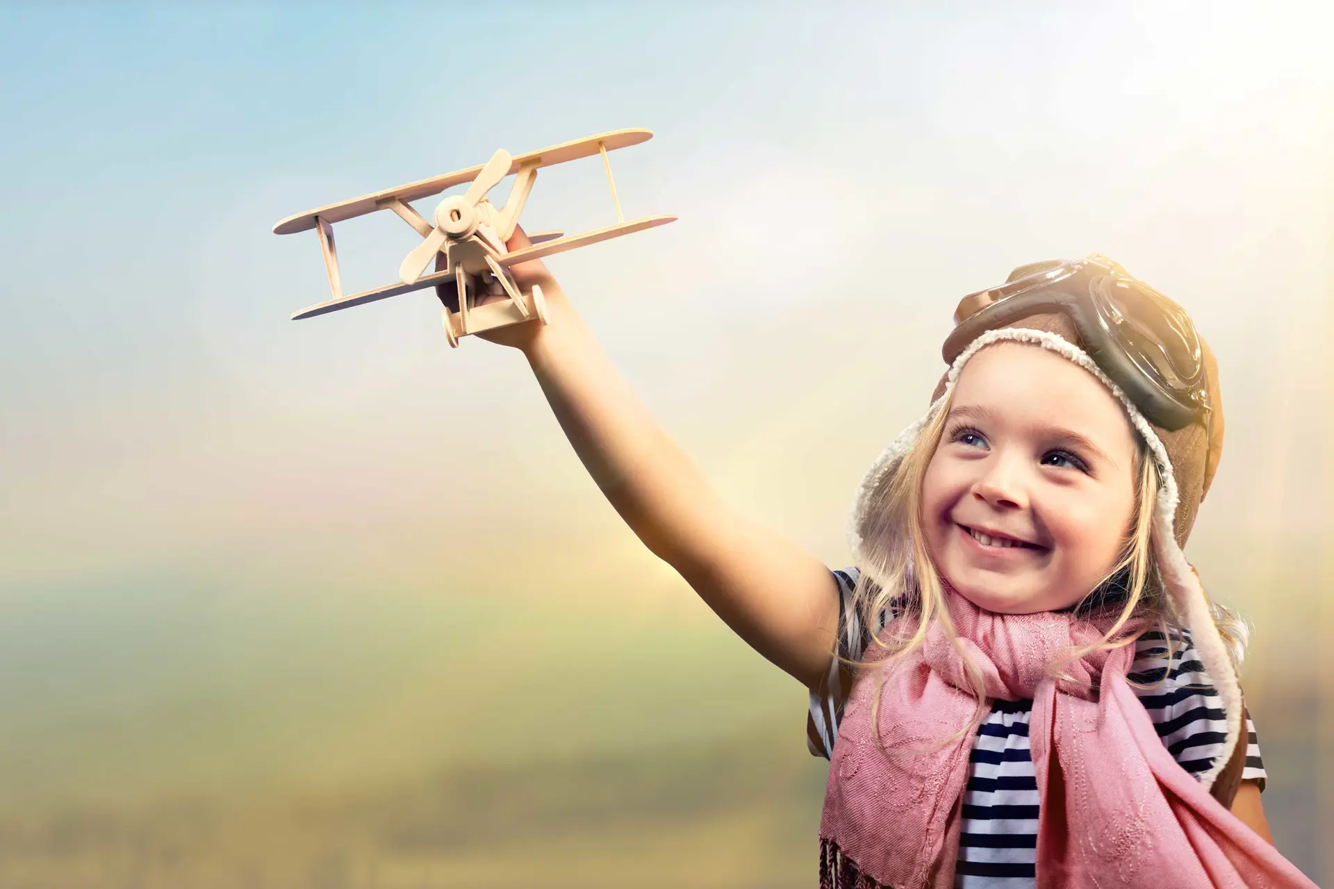 Lille pige med lyserødt tørklæde og flyvebriller holder et legetøjs fly i sin højre arm