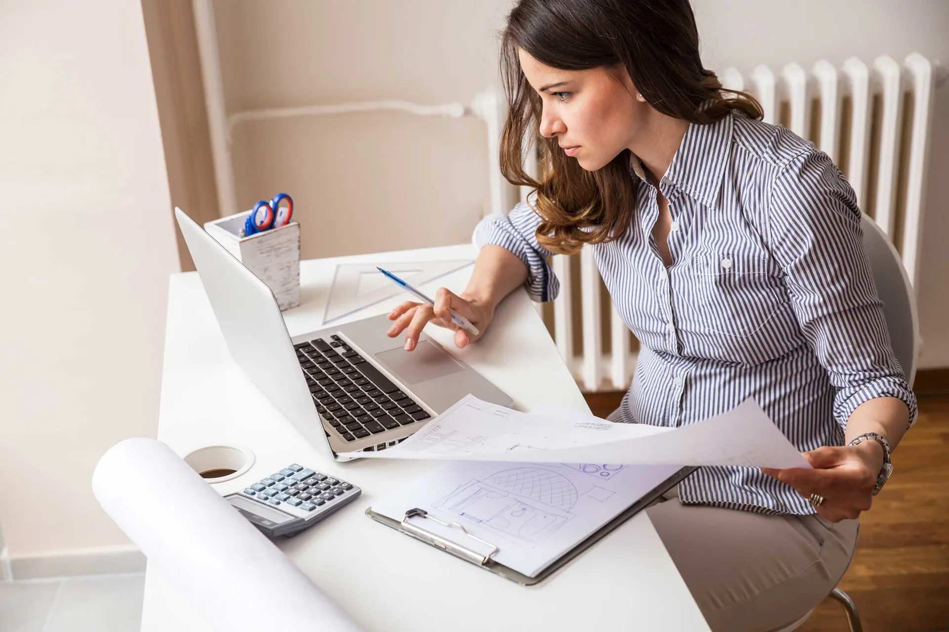 Dame sidder ved hvidt skrivebord med tegninger og computer. På bordet ligger en lineal, lommeregner og papir. 