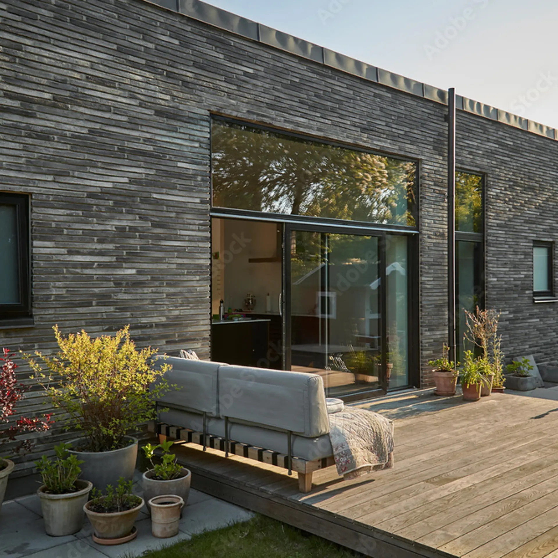 Sort murstenshus set udefra med træterrasse, udendørssofa, blomster i krukker og sorte vinduer.