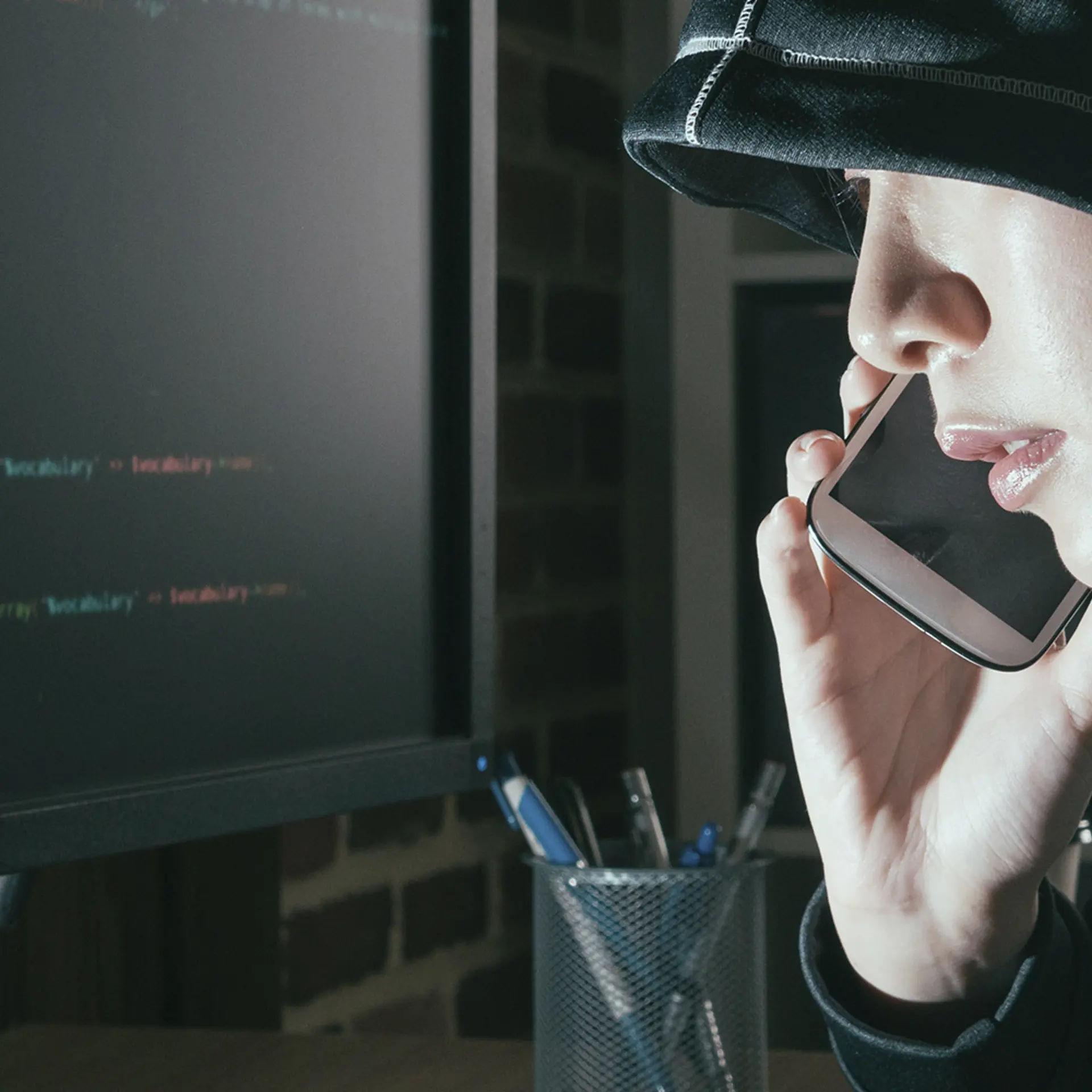 Hacker med sort hætte sidder foran computer med kodning, mens hackeren snakker i telefon