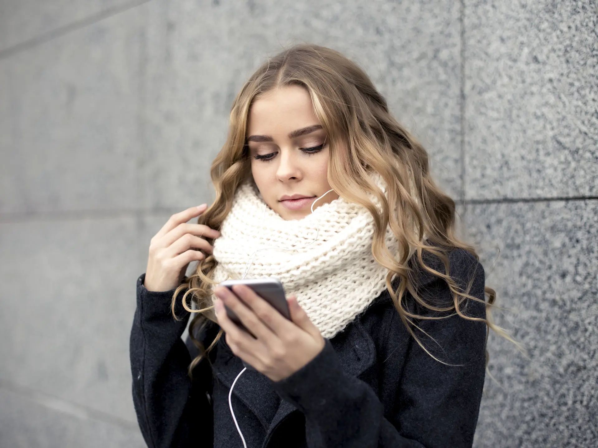 Ung pige med hvidt halstørklæde hører musik mens hun kigger på sin telefon 